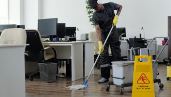 Contemporary young black man in workwear cleaning floor in openspace office Contemporary young black man in workwear cleaning floor in openspace office in front of yellow plastic signboard with caution