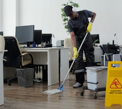 Contemporary young black man in workwear cleaning floor in openspace office in front of yellow plastic signboard with caution