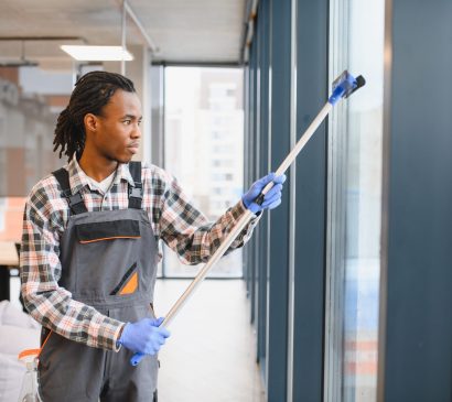 Skilled worker in uniform cleaning large office windows with a squeegee, ensuring clear and spotless glass surfaces in a modern workspace